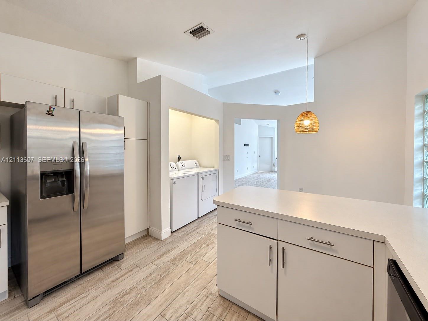 Kitchen featuring stainless steel side-by-side refrigerator, white cabinetry with silver-tone hardware, solid-surface countertops, wood-finish tile flooring, and glass block window detailing Kitchen featuring stainless steel side-by-side refrigerator, white cabinetry with silver-tone hardware, solid-surface countertops, wood-finish tile flooring, and glass block window detailing