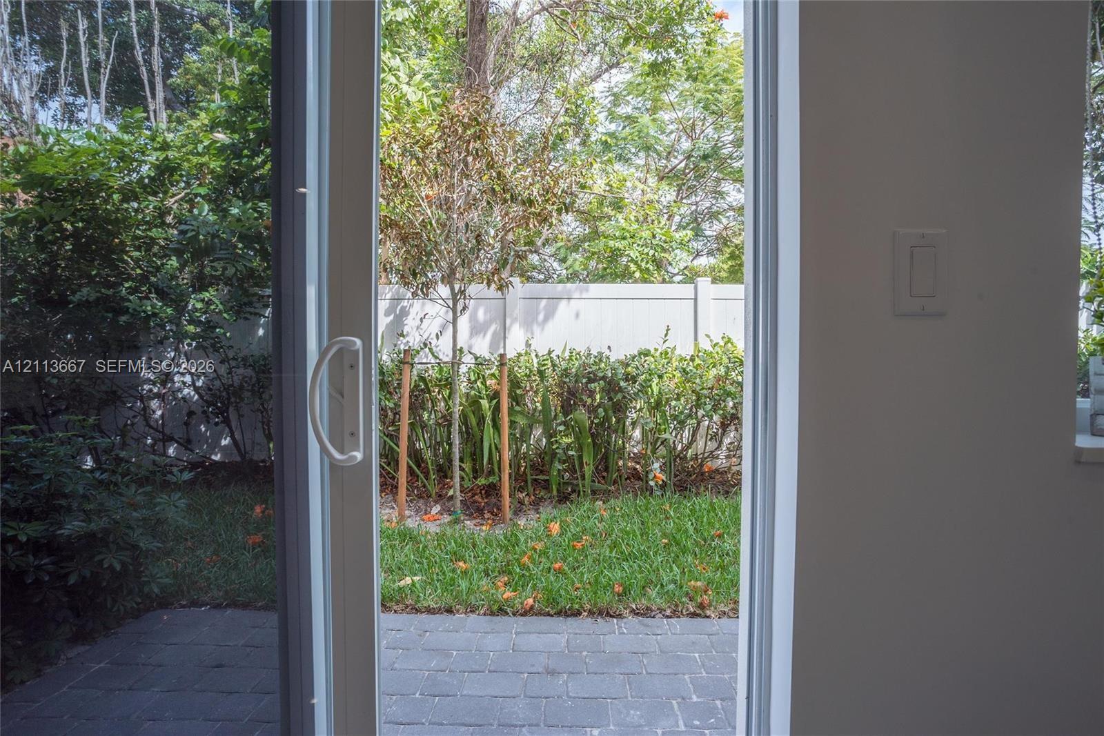 Sliding glass door opening to a paved patio, bordered by a lawn, mature shrubs, and a privacy fence Sliding glass door opening to a paved patio, bordered by a lawn, mature shrubs, and a privacy fence