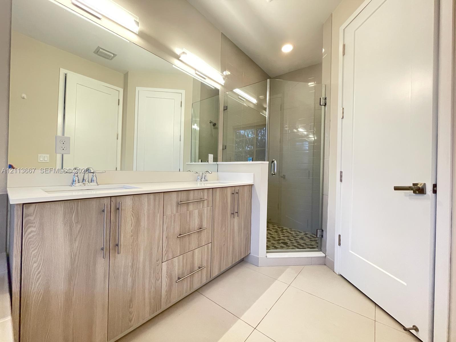 Dual-vanity cabinetry with wood-finish doors, solid-surface countertops, full-width mirror, and a glass-enclosed shower with tiled flooring