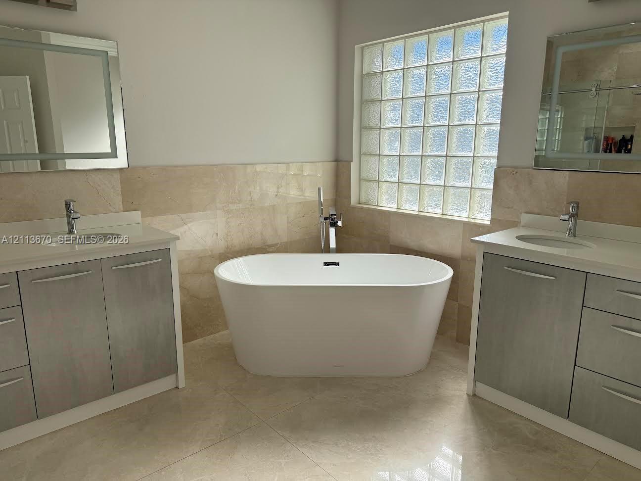 Freestanding oval soaking tub with floor-mounted faucet, dual vanities with integrated sinks, stone-finish tile backsplash, and glass block window