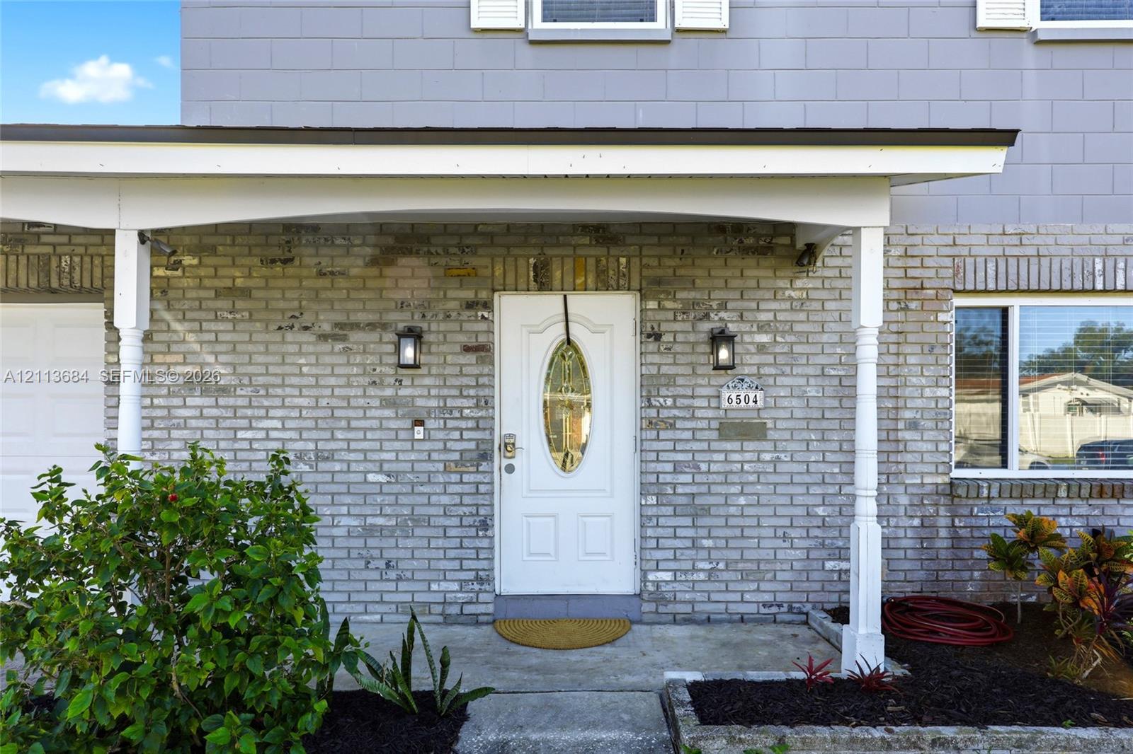 Exterior facade featuring light-toned brick masonry, a covered porch with spindle columns, and an entry door with an oval glass insert Exterior facade featuring light-toned brick masonry, a covered porch with spindle columns, and an entry door with an oval glass insert