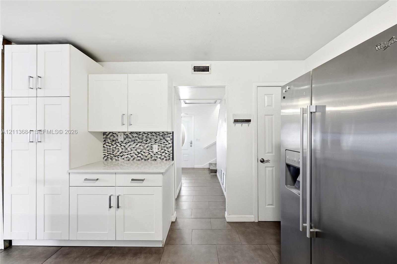 White shaker-style cabinetry with rectangular hardware, stone-composite countertop, geometric tile backsplash, large-format tile flooring, and stainless steel side-by-side refrigerator