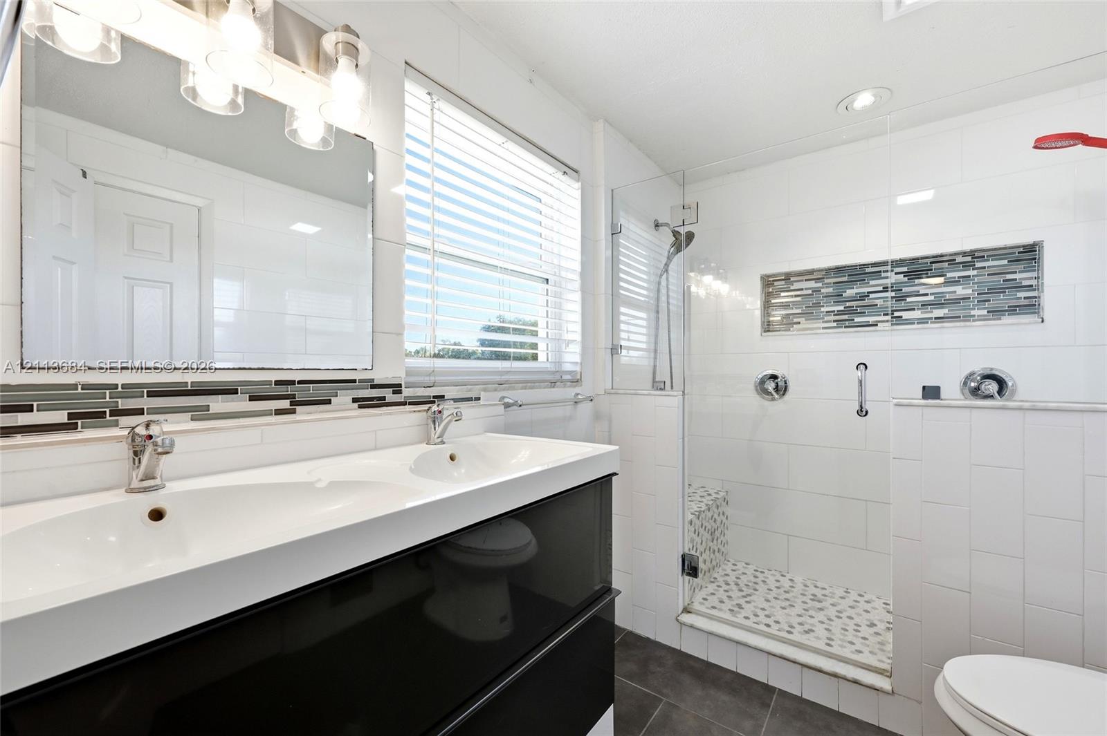 Dual-sink vanity with a white countertop and high-gloss cabinetry, glass-enclosed shower featuring stone tile flooring and decorative inlay, and white subway-style wall tiling