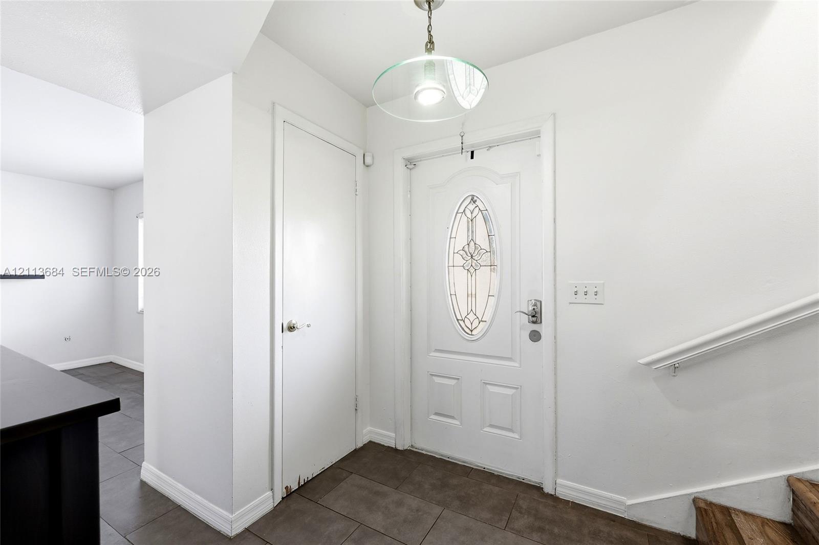 Entryway featuring tile flooring, a white exterior door with an oval glass insert, a pendant light fixture, and a wall-mounted handrail Entryway featuring tile flooring, a white exterior door with an oval glass insert, a pendant light fixture, and a wall-mounted handrail