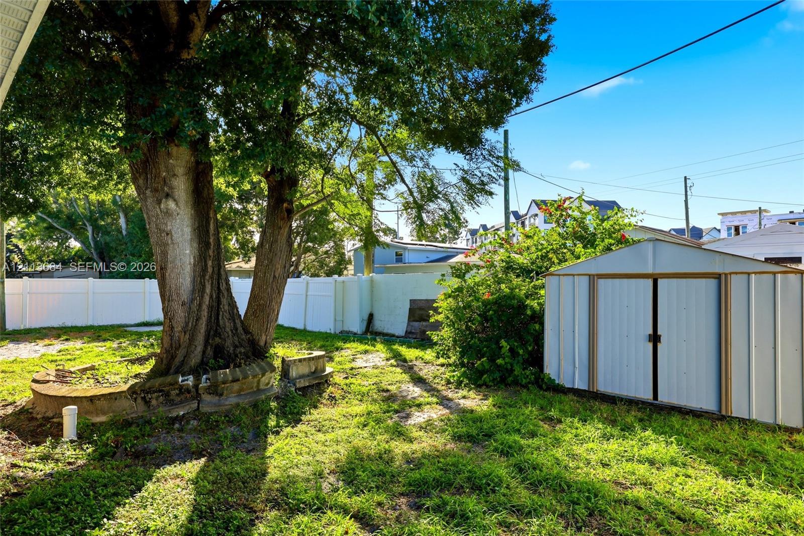 Backyard featuring a white vinyl privacy fence, mature shade tree with a stone-accented perimeter, and a detached metal storage structure