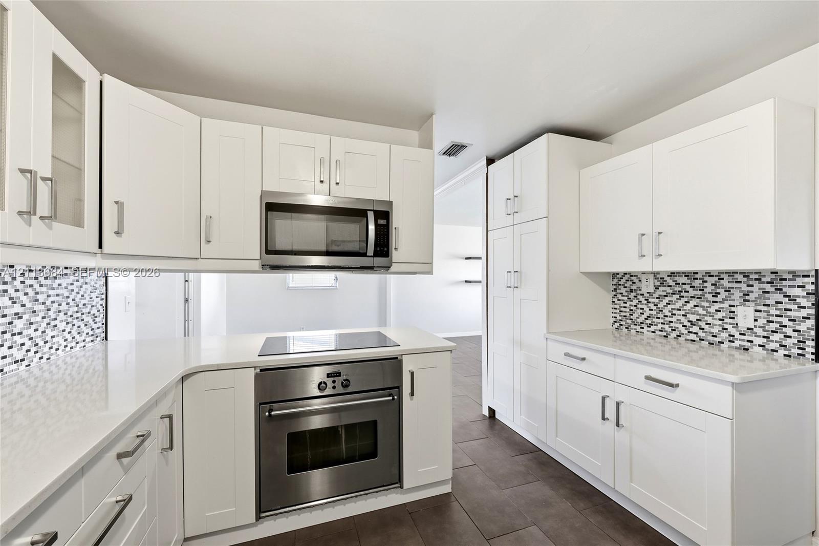 Kitchen featuring white shaker-style cabinetry, solid-surface countertops, a geometric mosaic tile backsplash, stainless steel built-in oven and microwave, and large-format tile flooring