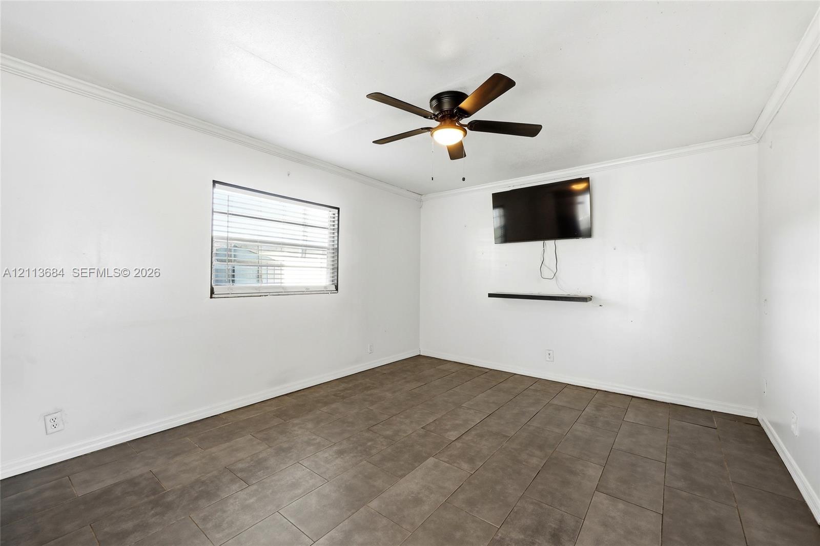 Neutral-toned room featuring large-format tile flooring, a dark-finish ceiling fan with integrated lighting, and a horizontal wall-mounted display shelf