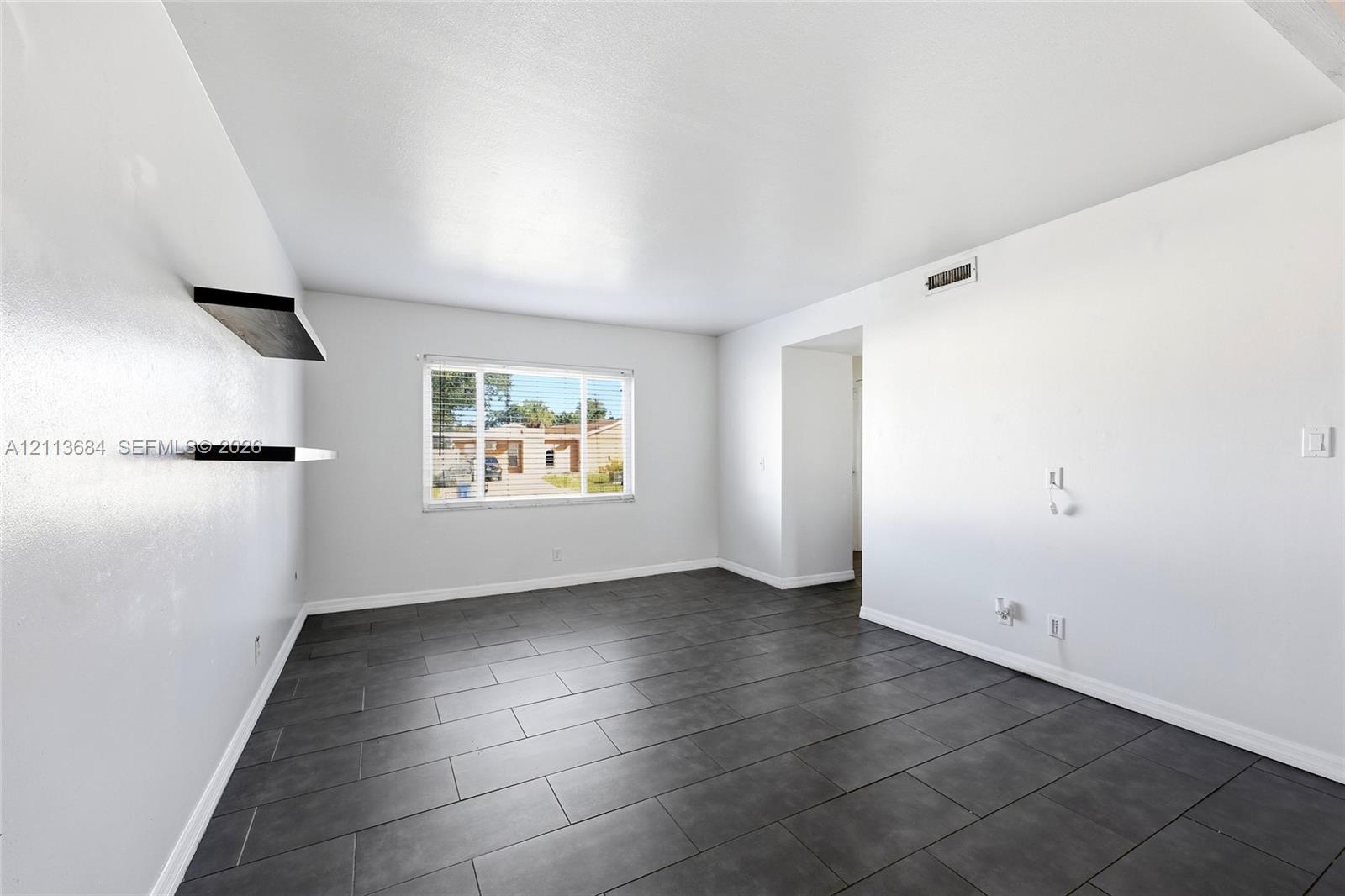 Interior space featuring large-format dark grey tile flooring, neutral wall finishes, a single-pane window, and two wall-mounted floating shelves Interior space featuring large-format dark grey tile flooring, neutral wall finishes, a single-pane window, and two wall-mounted floating shelves