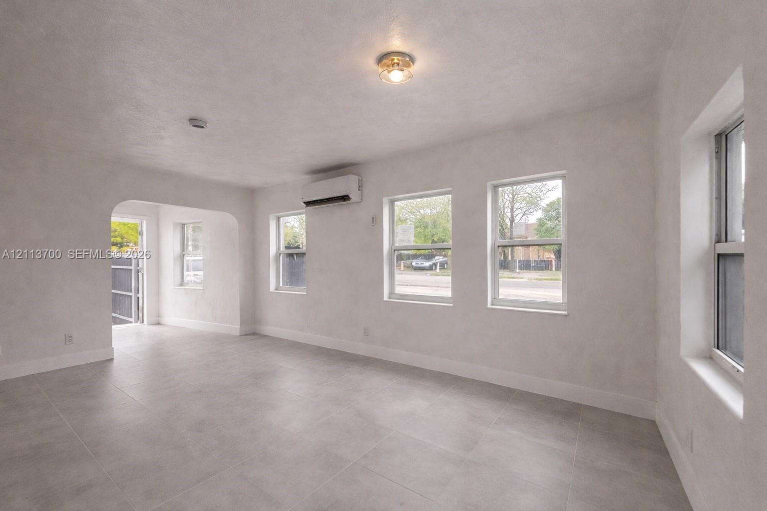 Neutral-toned tile flooring, smooth-finish walls, arched threshold, dual-pane windows, and wall-mounted climate control unit