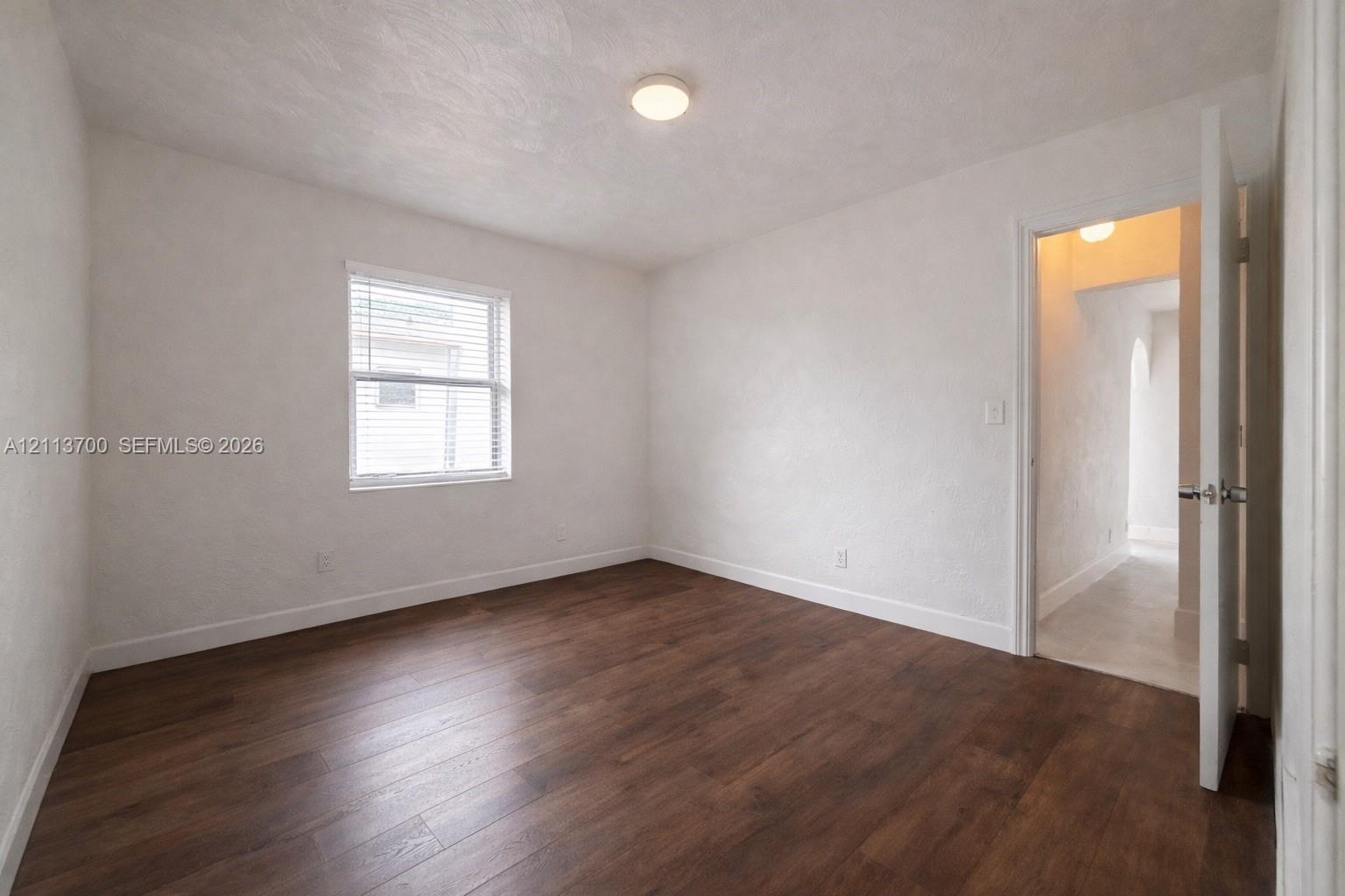 Interior room featuring wood-finish flooring, white textured walls, a single-hung window with horizontal blinds, and a flush-mount ceiling light