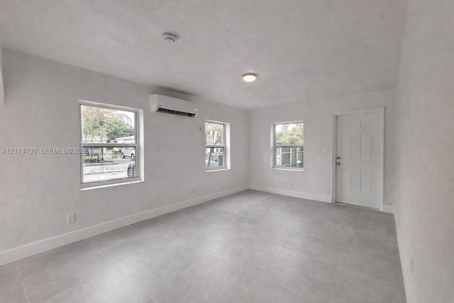 Neutral-toned tile flooring, three large windows, wall-mounted ductless climate control unit, and six-panel interior door Neutral-toned tile flooring, three large windows, wall-mounted ductless climate control unit, and six-panel interior door