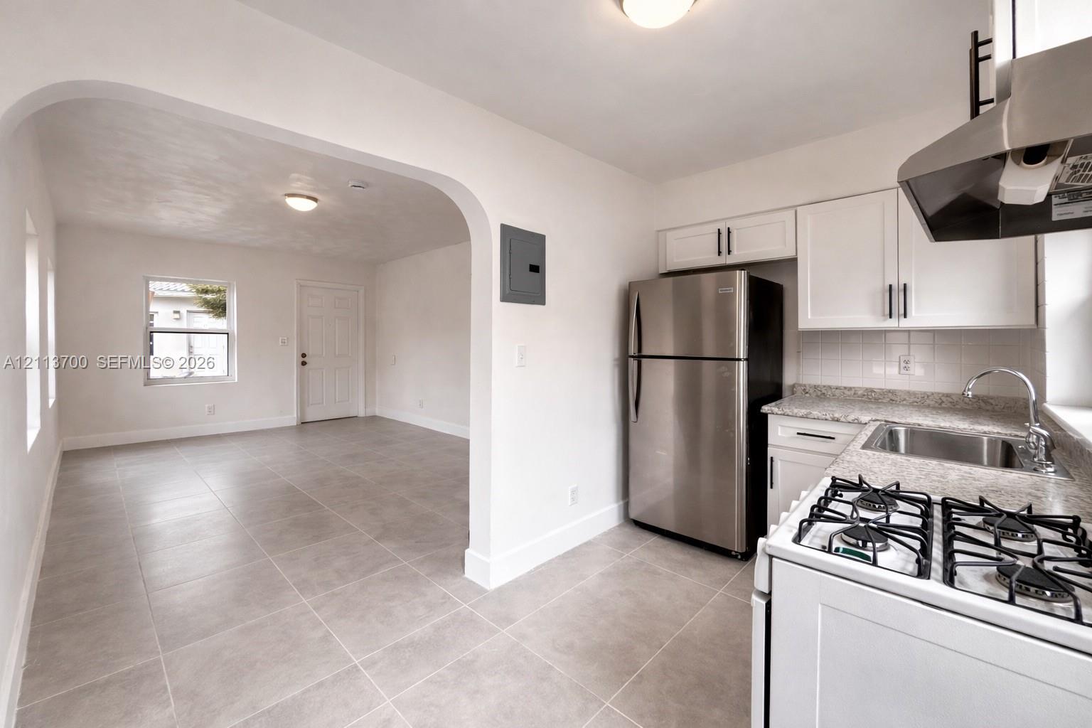 Kitchen featuring white Shaker-style cabinetry, stone-finish countertops, stainless steel appliances, and tile flooring, highlighted by an architectural arched wall opening Kitchen featuring white Shaker-style cabinetry, stone-finish countertops, stainless steel appliances, and tile flooring, highlighted by an architectural arched wall opening