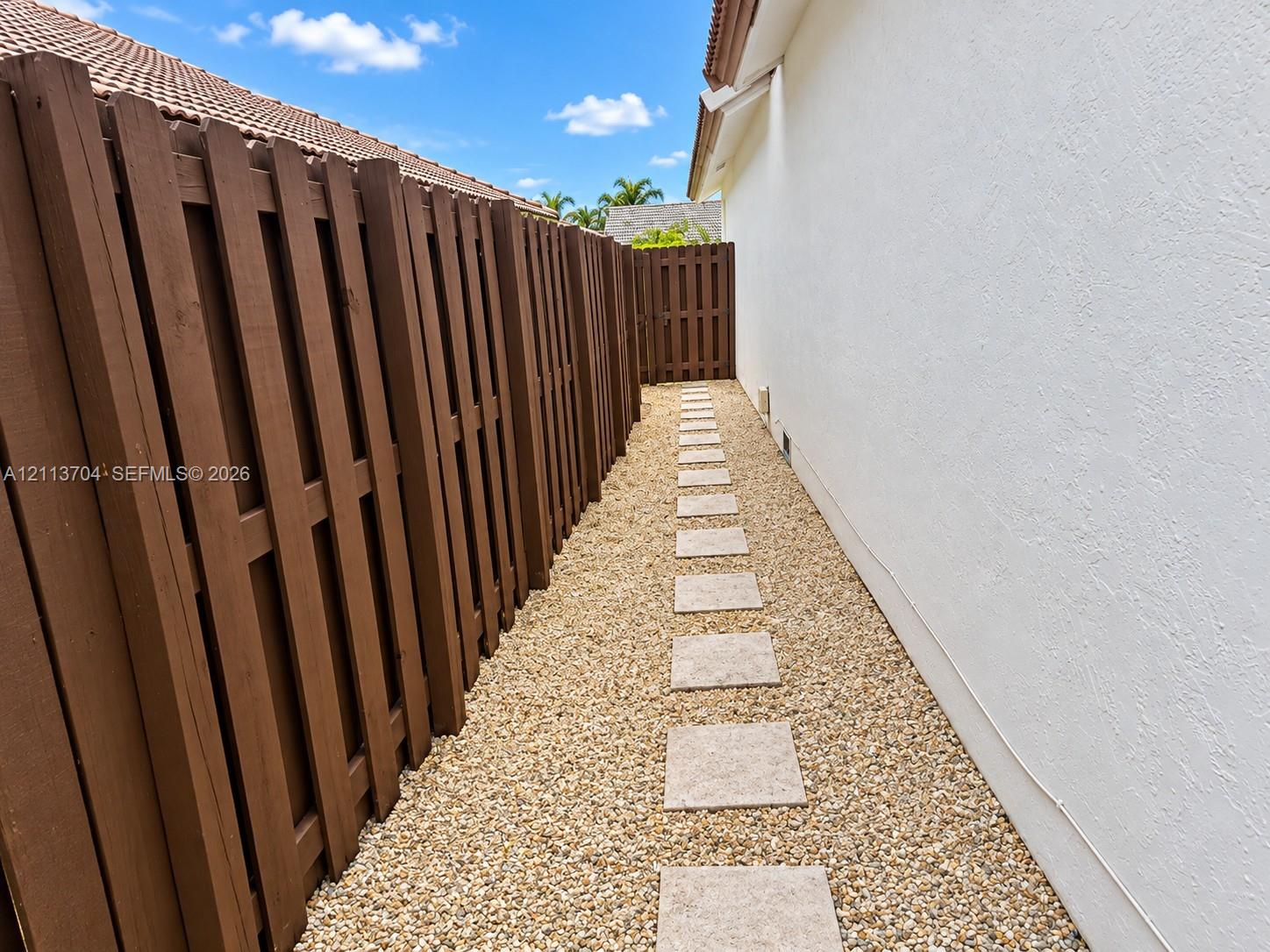 Exterior side yard featuring a white stucco wall, wood-finish privacy fencing, a decorative stone pathway, and a gravel ground surface