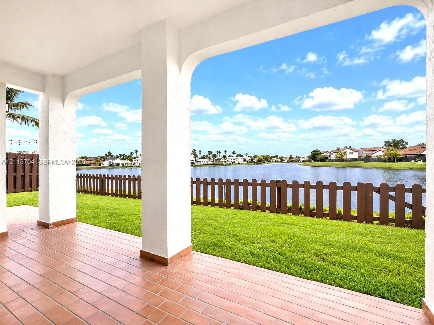 Covered patio featuring ceramic tile flooring, arched structural pillars, and direct water frontage with a wooden perimeter fence