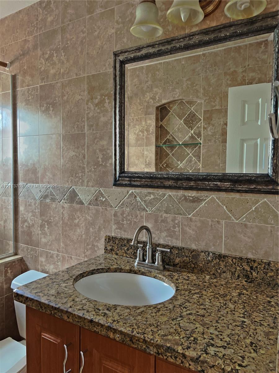 Vanity featuring a stone-finish countertop with an undermount sink, wood-finish cabinetry, full-height wall tiling with decorative tile inlay, and a built-in recessed wall niche