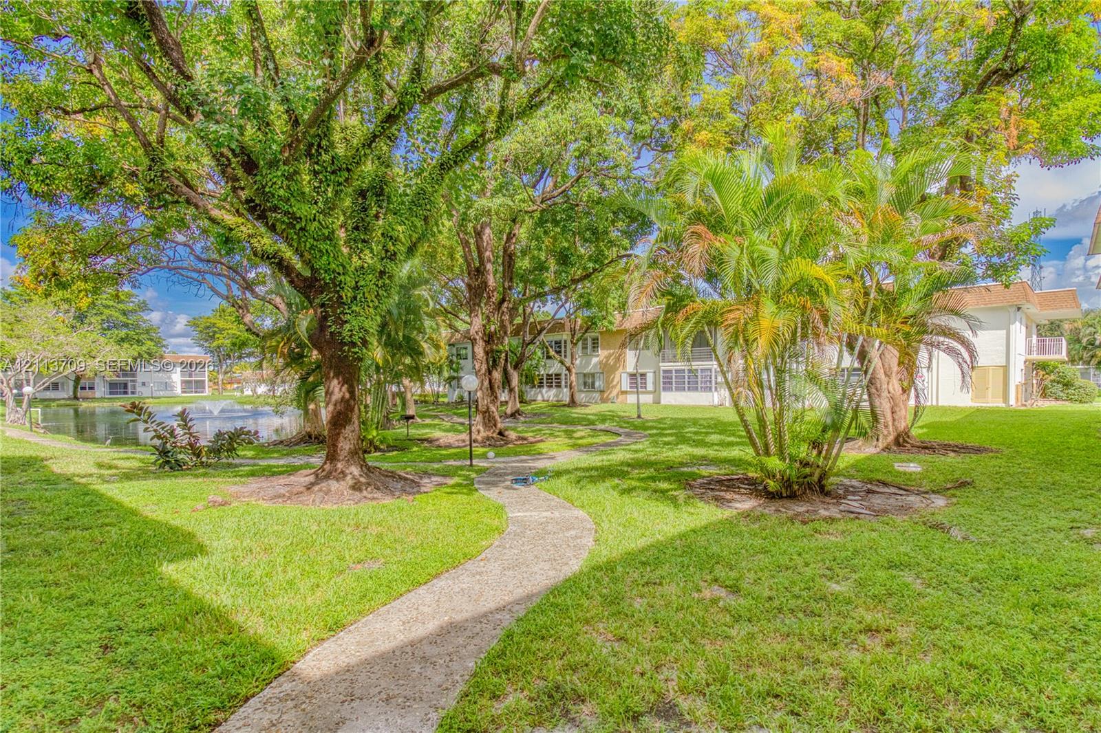 Landscaped grounds featuring a paved walking path, mature trees, ornamental palms, and a water feature