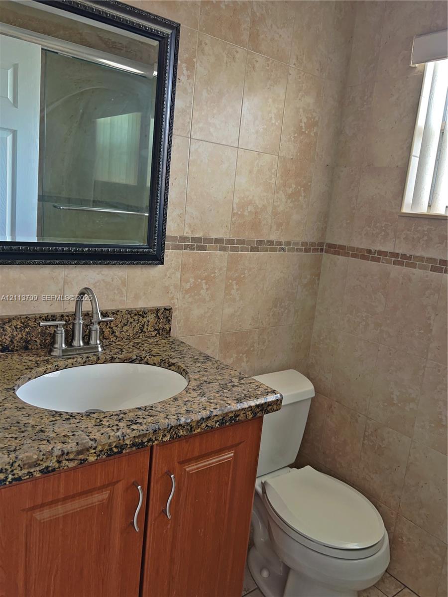 Bathroom vanity featuring a stone countertop with an undermount sink, wood-finish cabinetry, decorative wall tile with a mosaic accent border, and a wall-mounted framed mirror