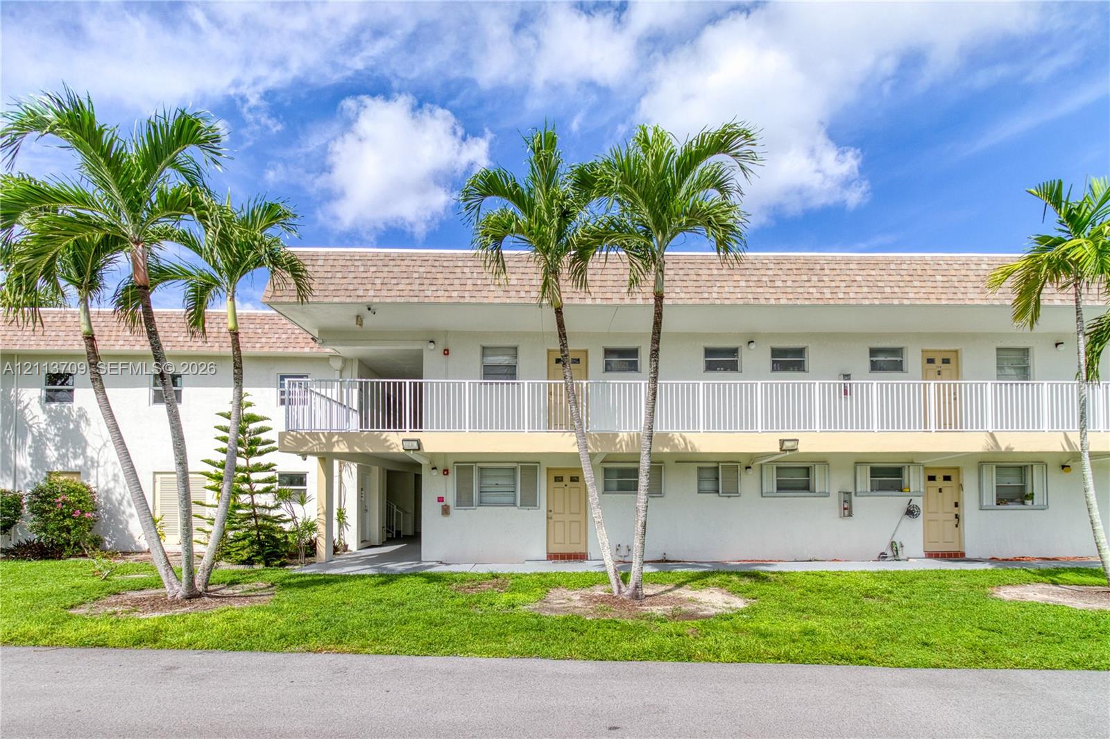 Two-story building exterior featuring a white stucco facade, shingled roof, white metal balcony railing, and mature palm trees