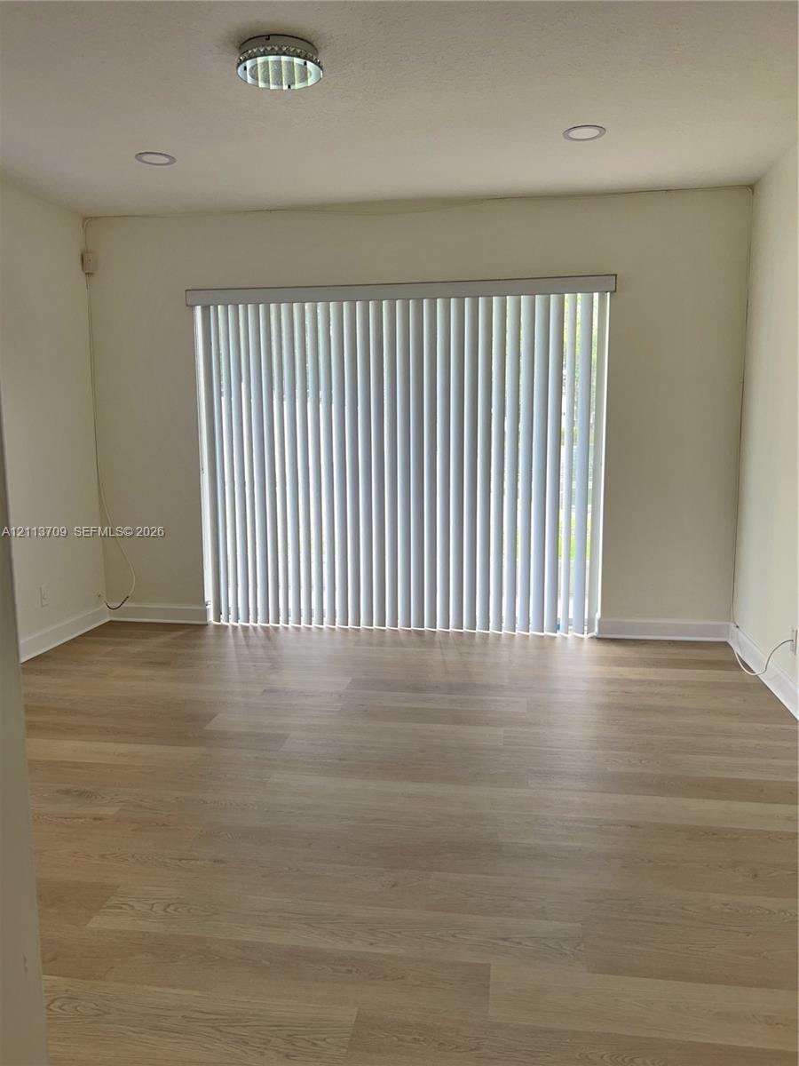 Neutral-toned room featuring wood-finish flooring, recessed lighting, and vertical window blinds Neutral-toned room featuring wood-finish flooring, recessed lighting, and vertical window blinds