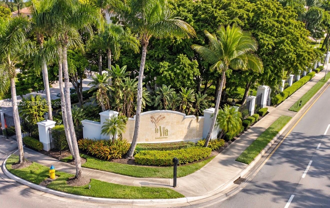 Community entrance monument featuring a stone-finish masonry sign, mature palm trees, manicured hedges, and ornate metal fencing