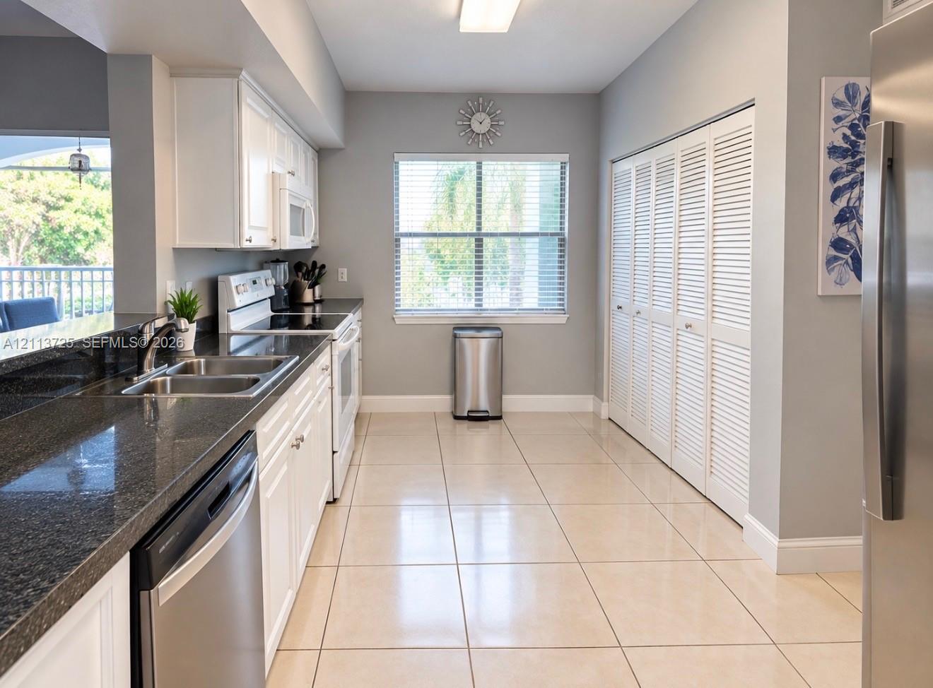 Kitchen featuring white shaker-style cabinetry, dark stone-finish countertops, stainless steel appliances, large-format tile flooring, and floor-to-ceiling louvered closet doors Kitchen featuring white shaker-style cabinetry, dark stone-finish countertops, stainless steel appliances, large-format tile flooring, and floor-to-ceiling louvered closet doors