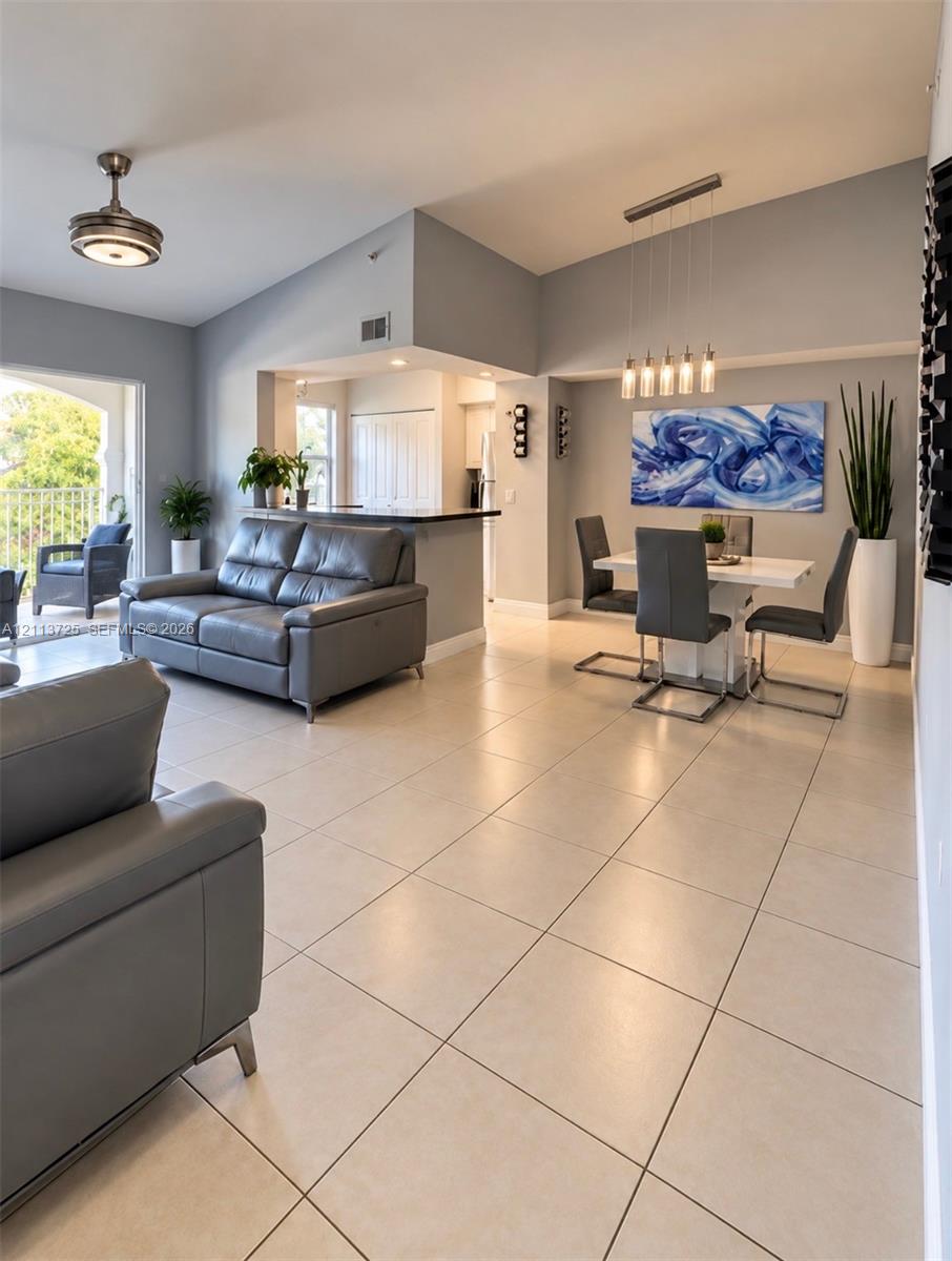 Open-concept interior featuring high vaulted ceilings, neutral large-format tile flooring, a breakfast bar with a dark countertop, and modern multi-pendant lighting Open-concept interior featuring high vaulted ceilings, neutral large-format tile flooring, a breakfast bar with a dark countertop, and modern multi-pendant lighting