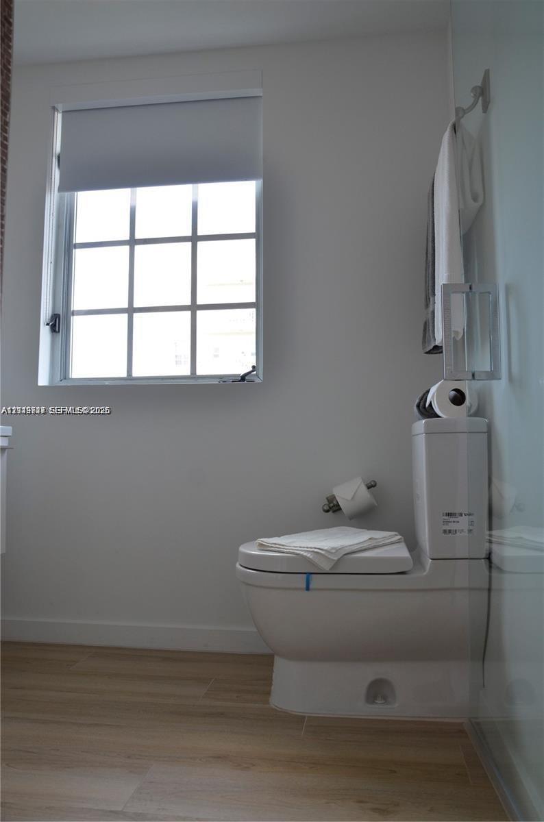 Bathroom featuring a dual-flush toilet, wood-finish flooring, multi-pane window with a roller shade, and wall-mounted hardware
