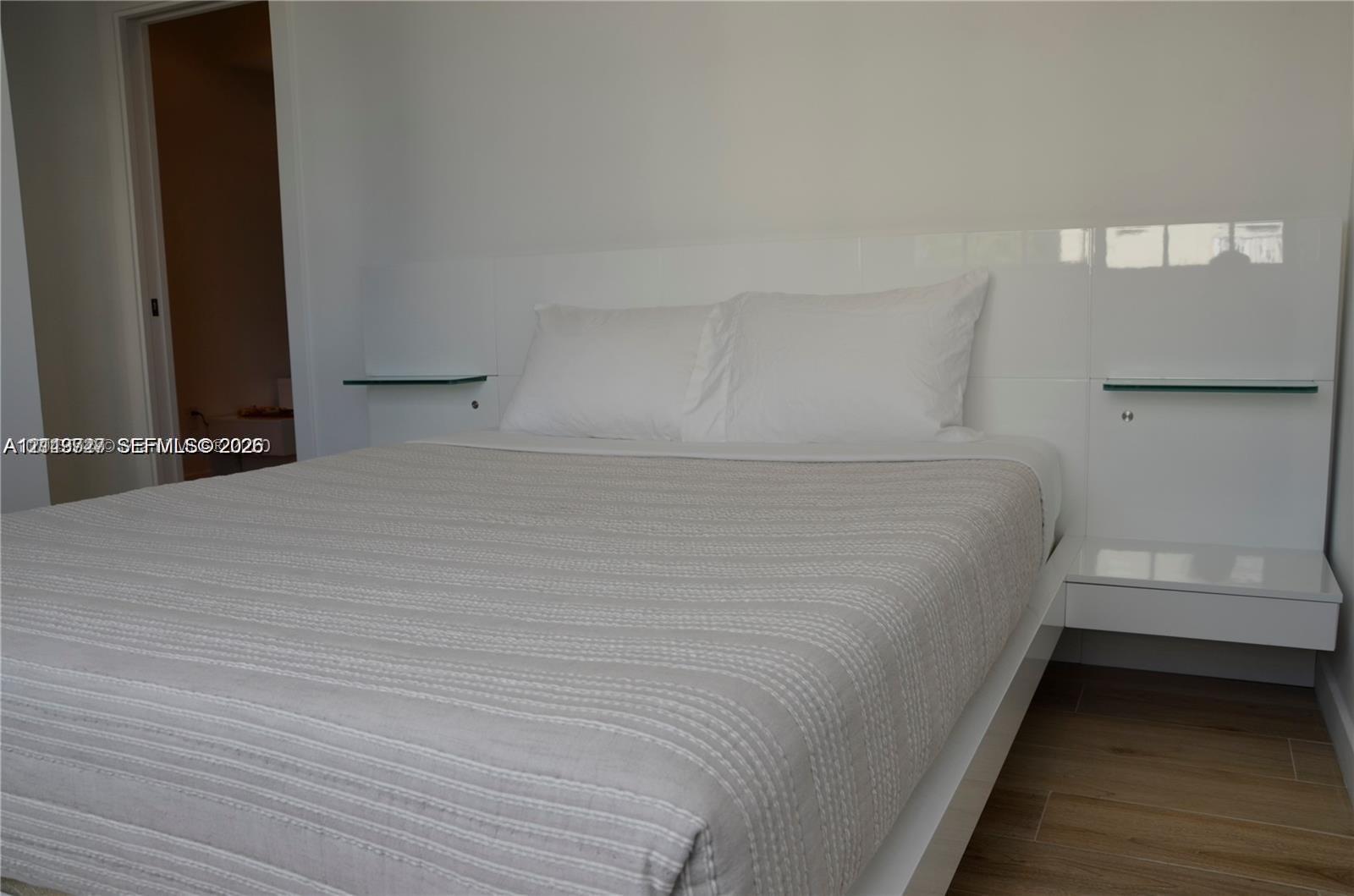 Built-in high-gloss white headboard featuring integrated glass shelving and floating side tables, complemented by wood-finish flooring