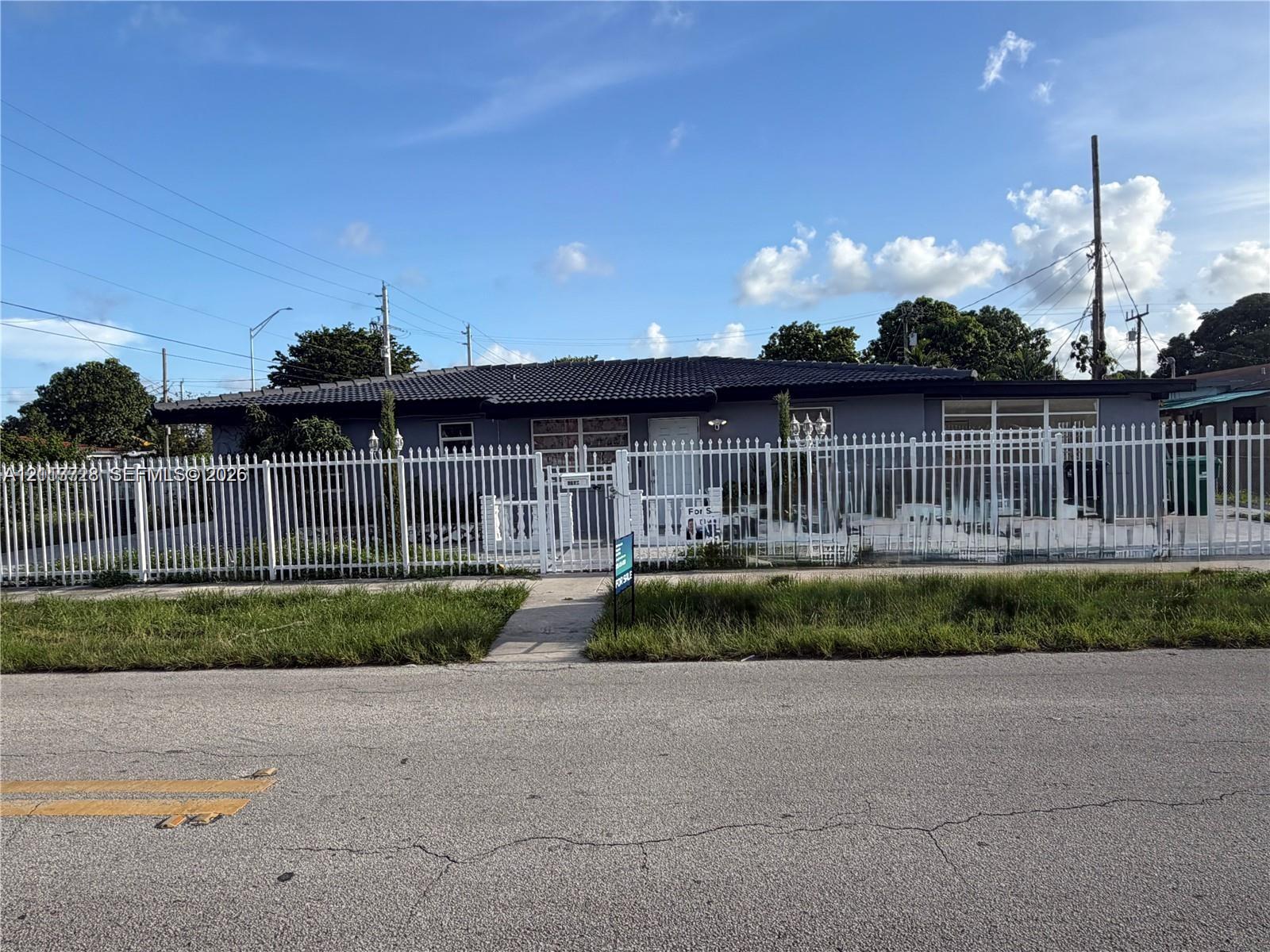 Single-story exterior featuring a dark-toned hipped roof, a white metal perimeter fence with an integrated pedestrian gate, and a concrete paver walkway