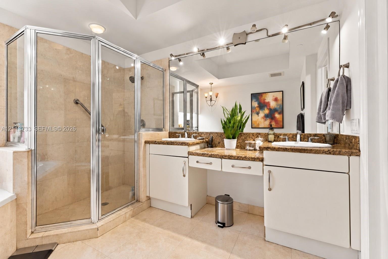 Double vanity featuring stone countertops with dual undermount sinks, central dressing table, full-width mirror with integrated track lighting, and a walk-in glass-enclosed shower with tile surround