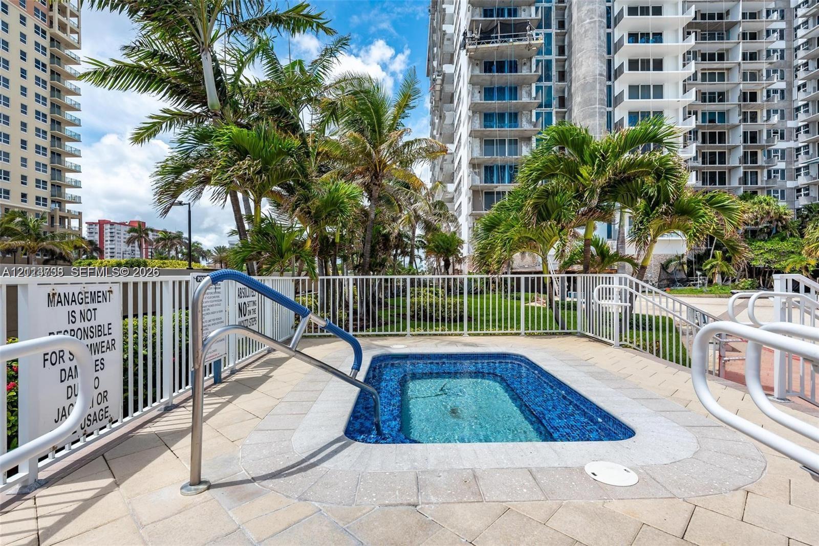 In-ground spa with tile lining, surrounded by a paved deck, white metal perimeter fencing, and mature palm trees