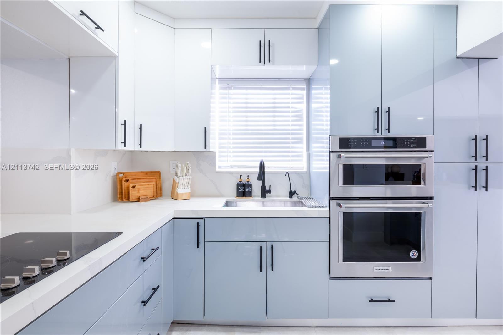 High-gloss cabinetry featuring white and pale blue finishes, solid surface countertops with integrated backsplash, built-in stainless steel double wall ovens, black hardware, and an undermount sink with a high-arch matte black faucet