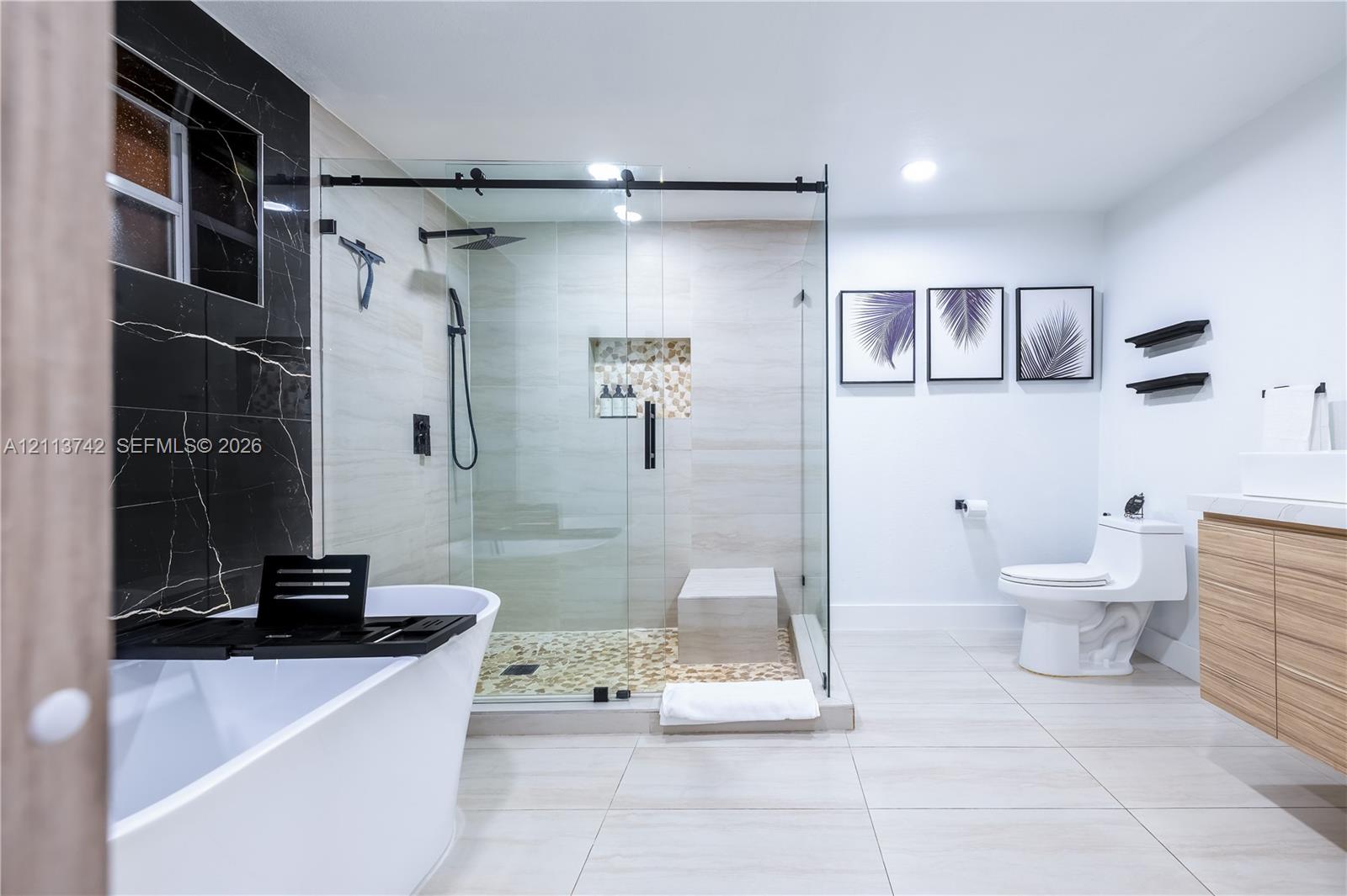 Freestanding soaking tub, glass-enclosed walk-in shower with stone tile flooring, built-in shower niche, wall-mounted wood-finish vanity, and floor-to-ceiling contrasting wall tile