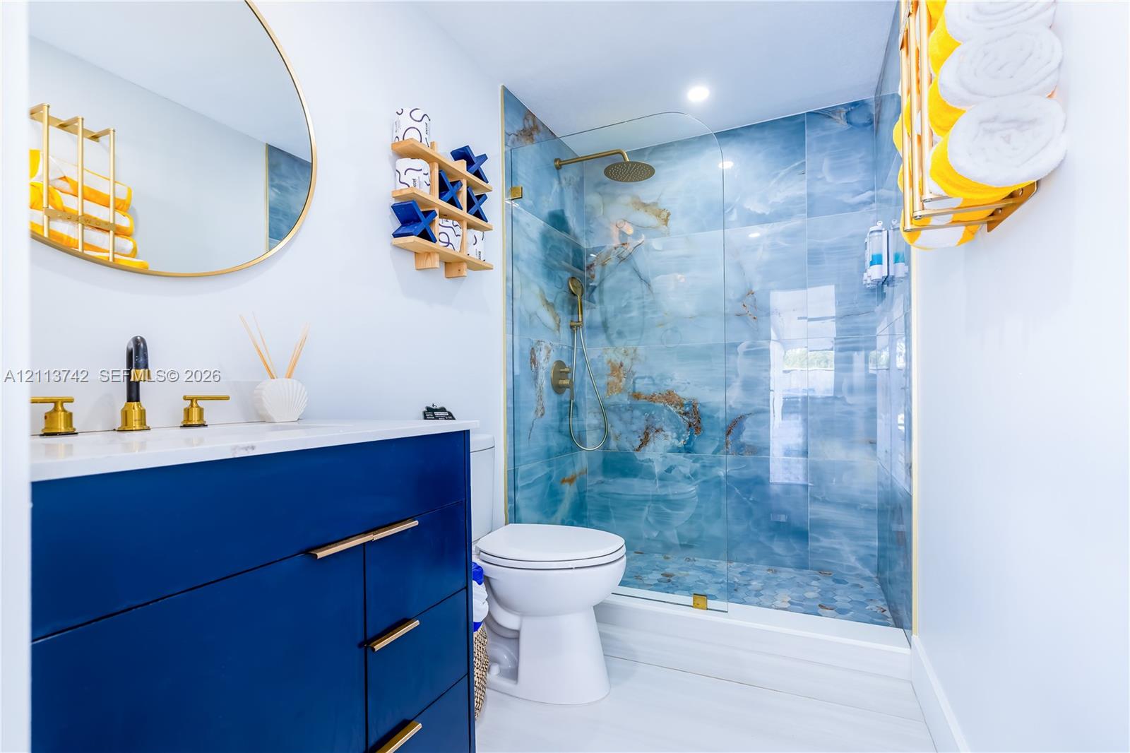 Blue vanity featuring a white countertop and gold-tone hardware, oval gold-framed mirror, and a walk-in shower with floor-to-ceiling patterned wall tile, a frameless glass door, and a gold-tone rainfall showerhead