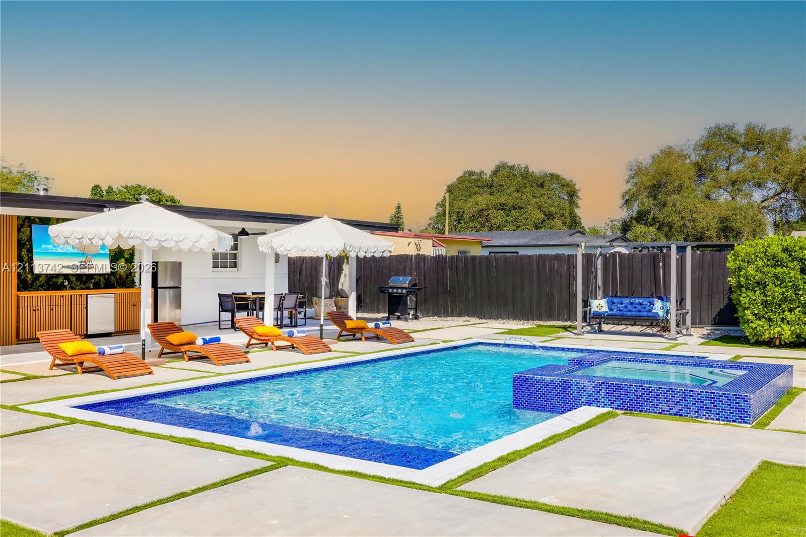 Backyard featuring a swimming pool with a raised tiled spa, concrete paver decking with grass inserts, and a covered outdoor kitchen with built-in cabinetry Backyard featuring a swimming pool with a raised tiled spa, concrete paver decking with grass inserts, and a covered outdoor kitchen with built-in cabinetry