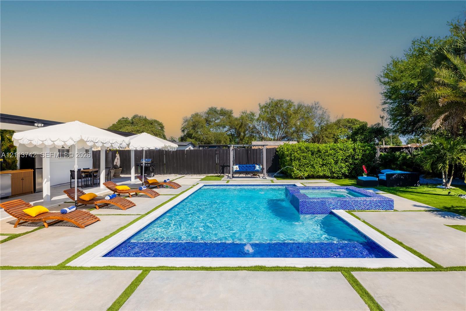 In-ground swimming pool featuring a tiled spa, concrete paver decking, integrated turf ribbons, and a covered outdoor kitchen area