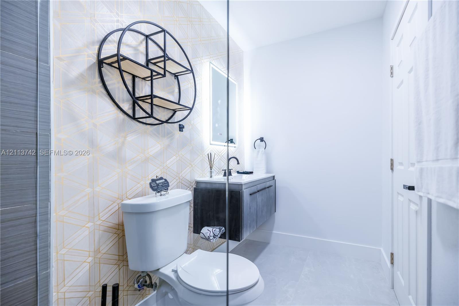 Modern floating vanity with a dark wood-finish cabinet, geometric accent wall, illuminated mirror, and matte black hardware