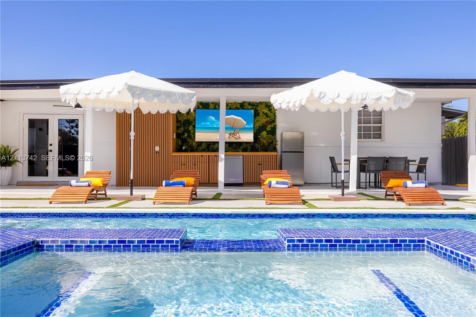 Outdoor swimming pool with blue tile edging, paired with a covered patio featuring built-in wood-slat cabinetry and a recessed television mounting area Outdoor swimming pool with blue tile edging, paired with a covered patio featuring built-in wood-slat cabinetry and a recessed television mounting area