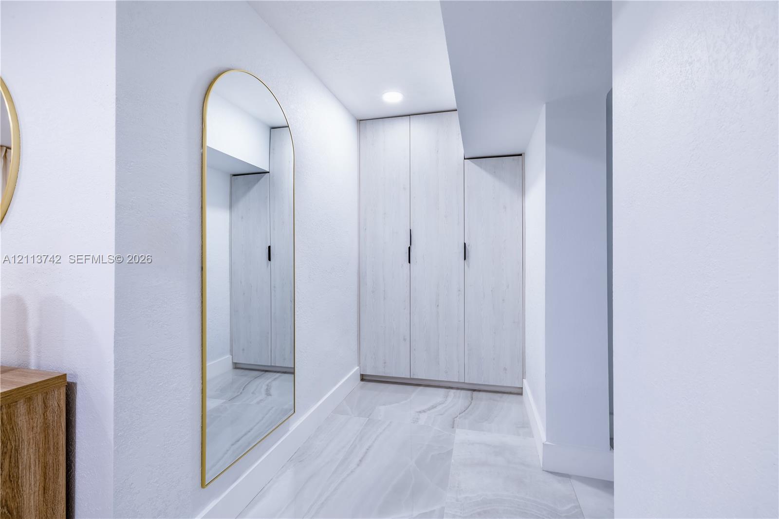 Built-in cabinetry with light-toned finish, full-length arched wall mirror with metallic frame, recessed ceiling lighting, and large-format porcelain tile flooring