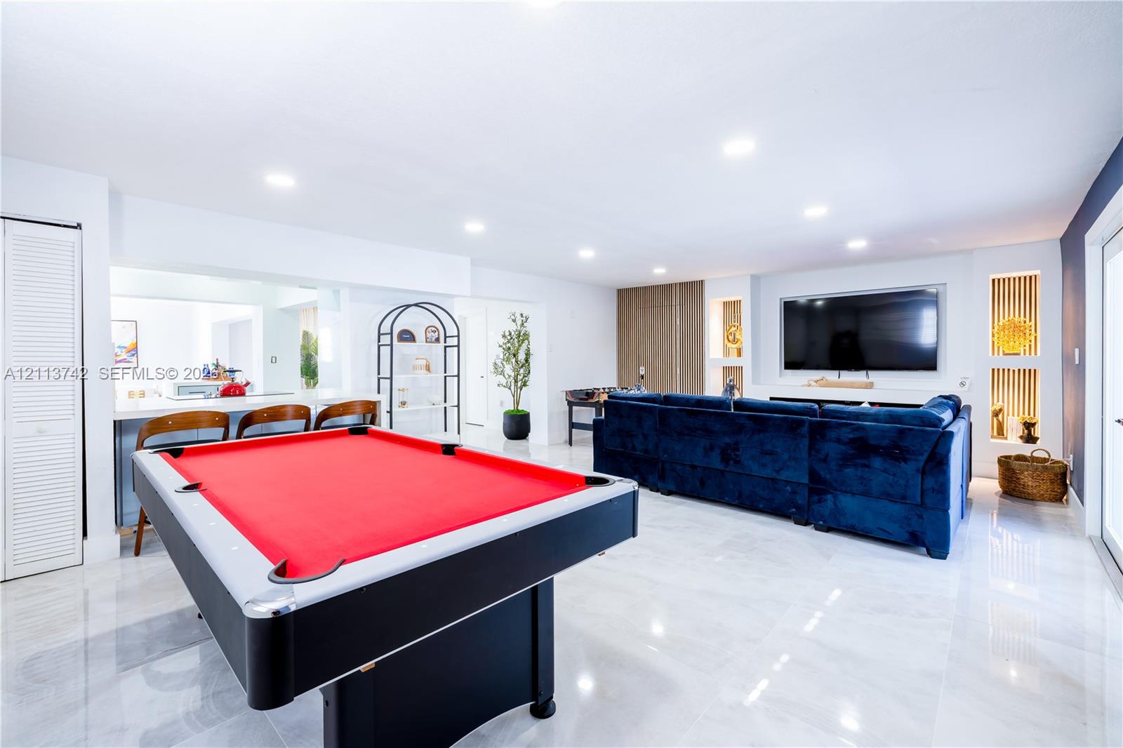 Large open-concept space featuring polished tile flooring, recessed ceiling lighting, a built-in media wall with integrated shelving, and a bar area with custom cabinetry