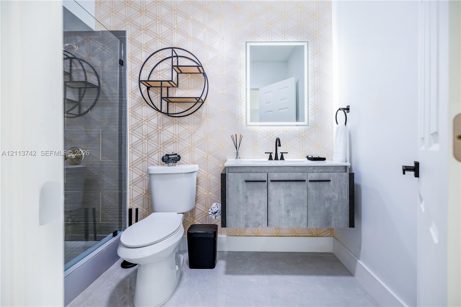 Floating vanity with a concrete-finish cabinet and solid surface countertop, geometric patterned wall covering, glass-enclosed shower with tiled surround, and LED-backlit mirror