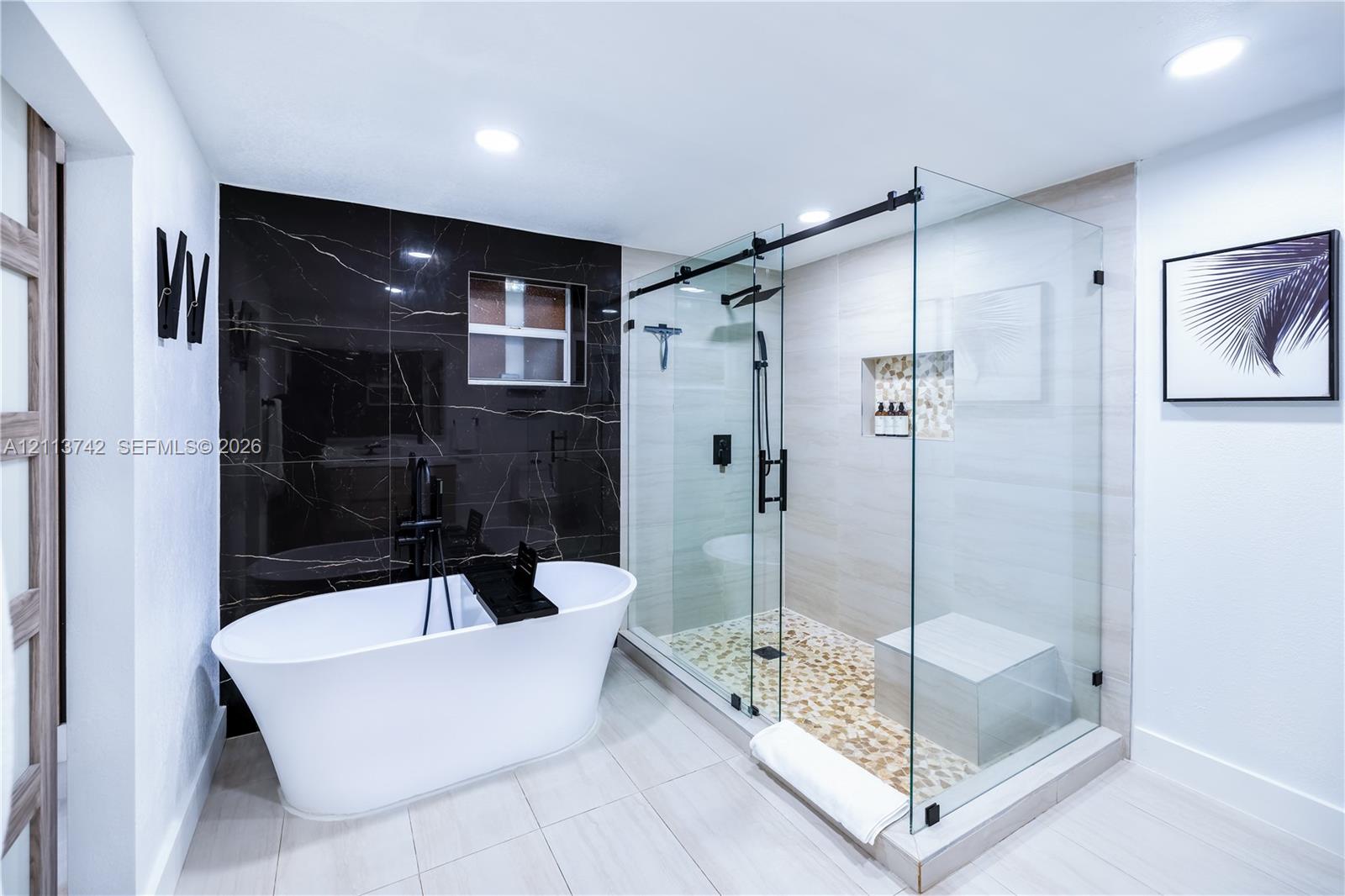 Freestanding soaking tub with matte black floor-mounted faucet, walk-in glass-enclosed shower with built-in bench, pebble-style shower flooring, recessed wall niche, and contrasting black tile accent wall Freestanding soaking tub with matte black floor-mounted faucet, walk-in glass-enclosed shower with built-in bench, pebble-style shower flooring, recessed wall niche, and contrasting black tile accent wall