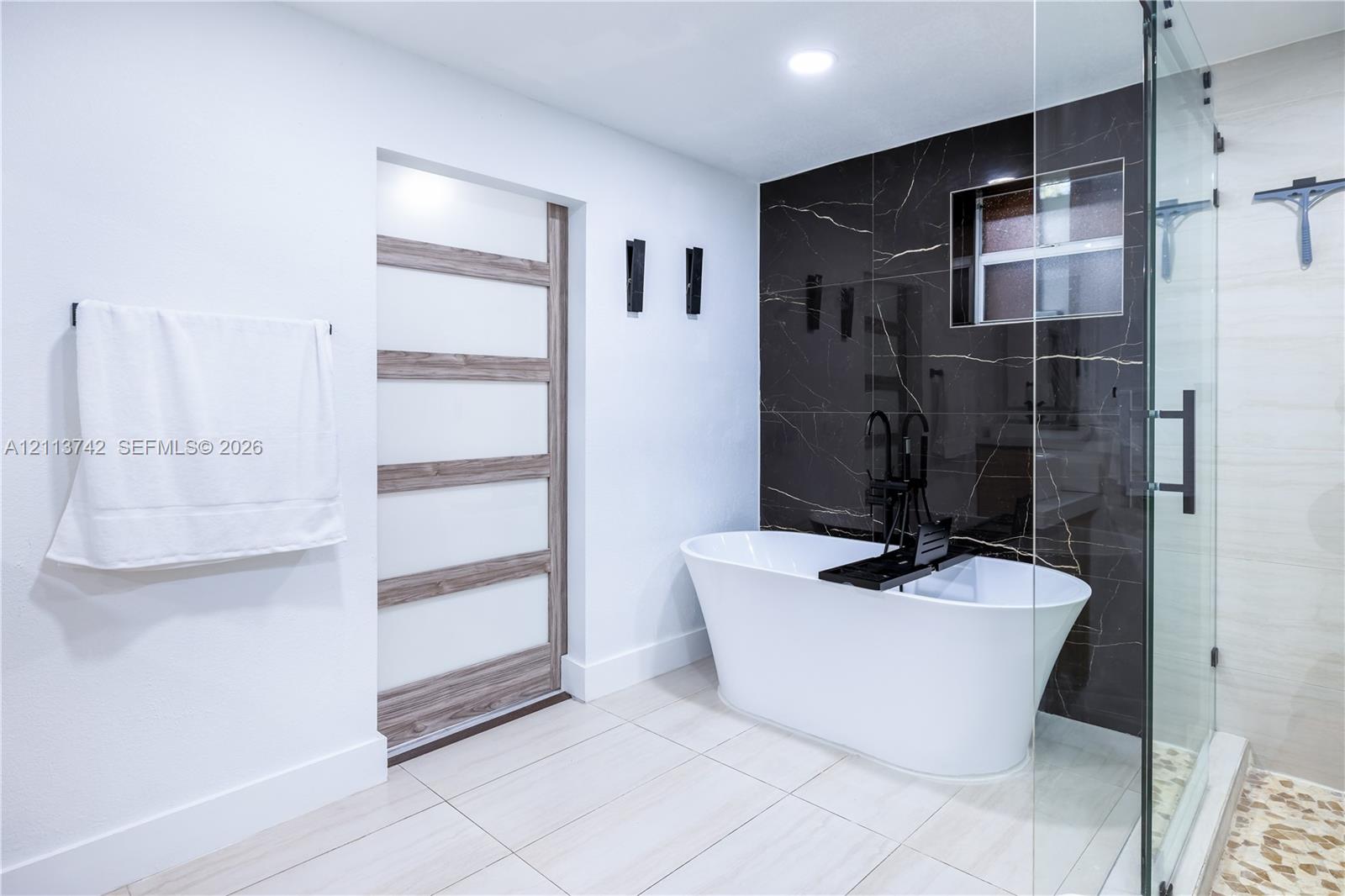 Freestanding soaking tub with matte black floor-mounted faucet, dark stone-finish accent wall, glass-enclosed shower, and multi-panel door with wood-finish detailing