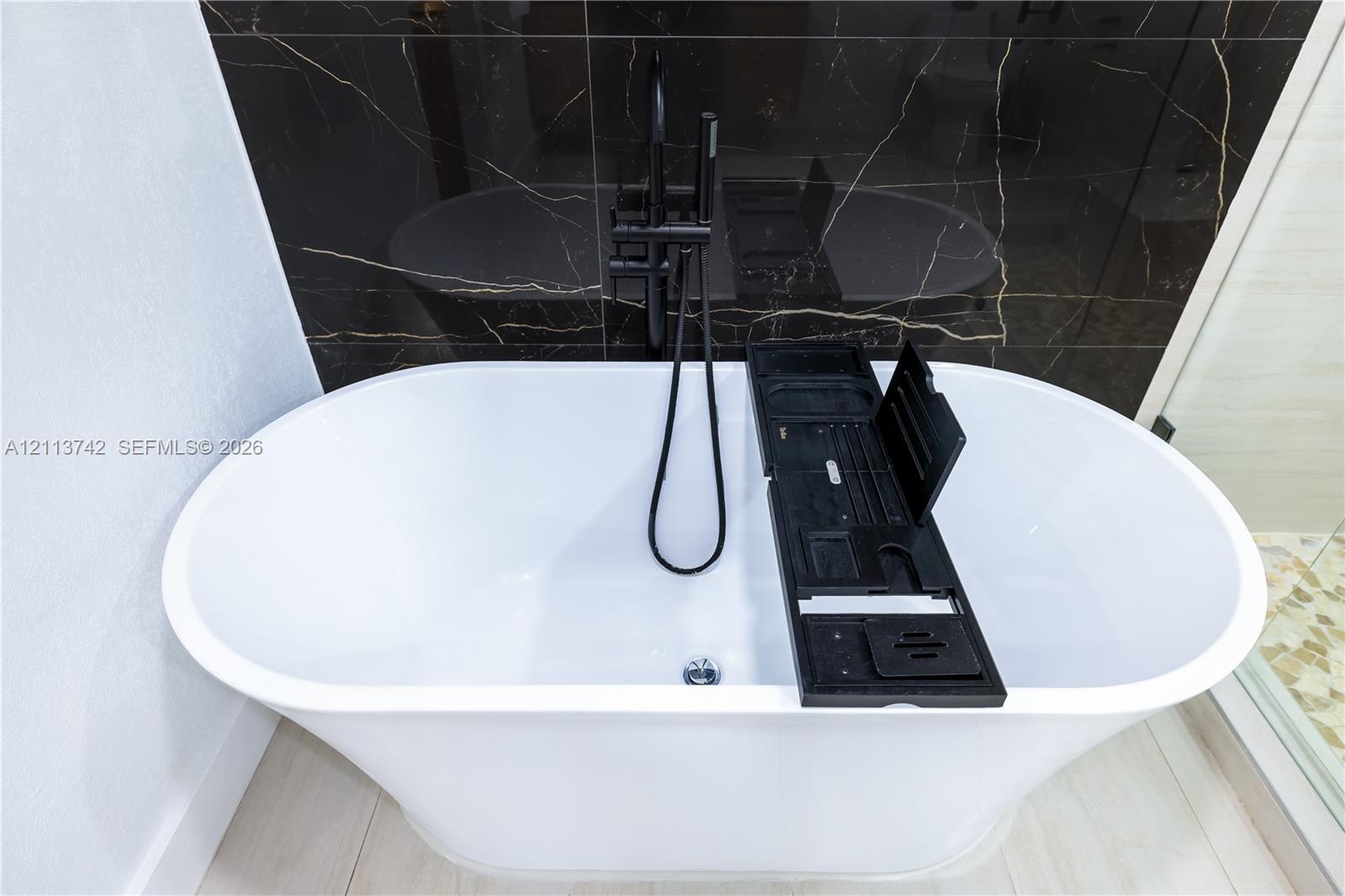 Freestanding white soaking tub with a floor-mounted matte black faucet and handheld shower attachment, set against a dark, veined tile accent wall