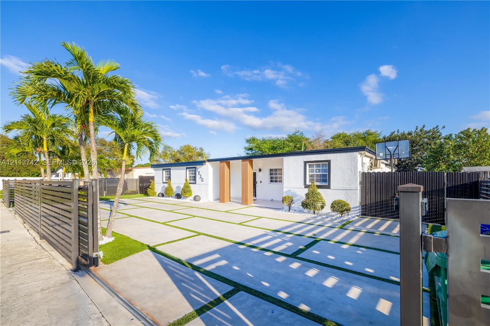Single-story exterior featuring white stucco siding, a concrete paver driveway with turf accents, mature palm trees, and perimeter horizontal slat fencing Single-story exterior featuring white stucco siding, a concrete paver driveway with turf accents, mature palm trees, and perimeter horizontal slat fencing