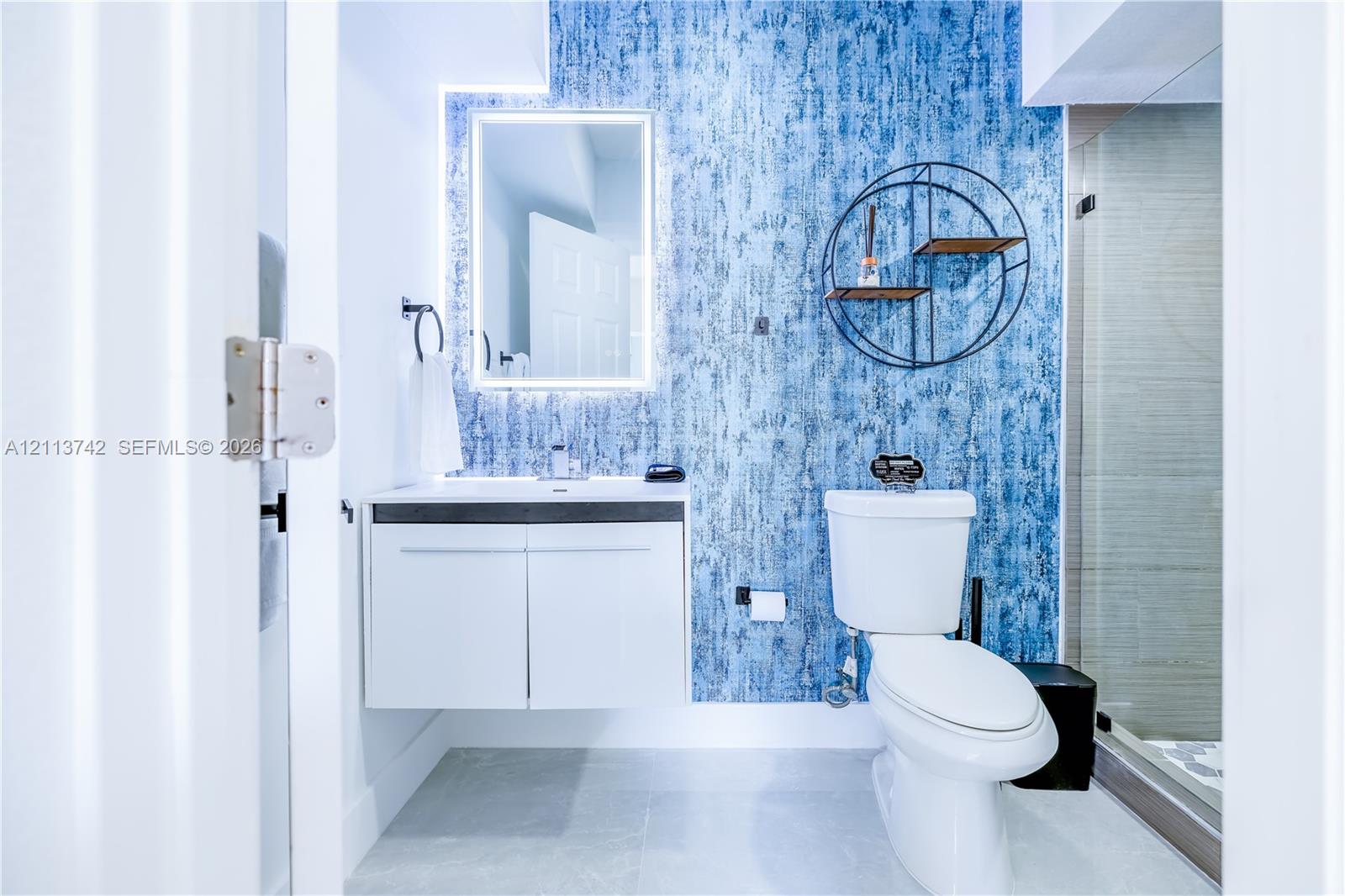 Bathroom featuring a wall-mounted floating vanity, backlit rectangular mirror, textured blue accent wall, and glass-enclosed shower