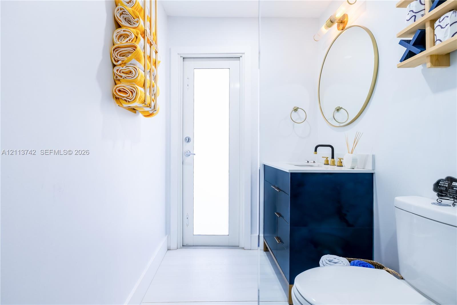 Bathroom featuring a dark blue vanity with gold hardware, an oval mirror with a gold frame, a modern wall-mounted light fixture, and a door with an inset glass panel