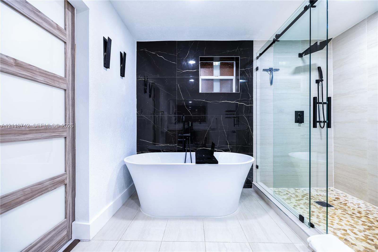 Freestanding soaking tub, walk-in glass-enclosed shower with pebble-style floor tiling, dark stone-finish accent wall, and wood-finish door with frosted glass panels