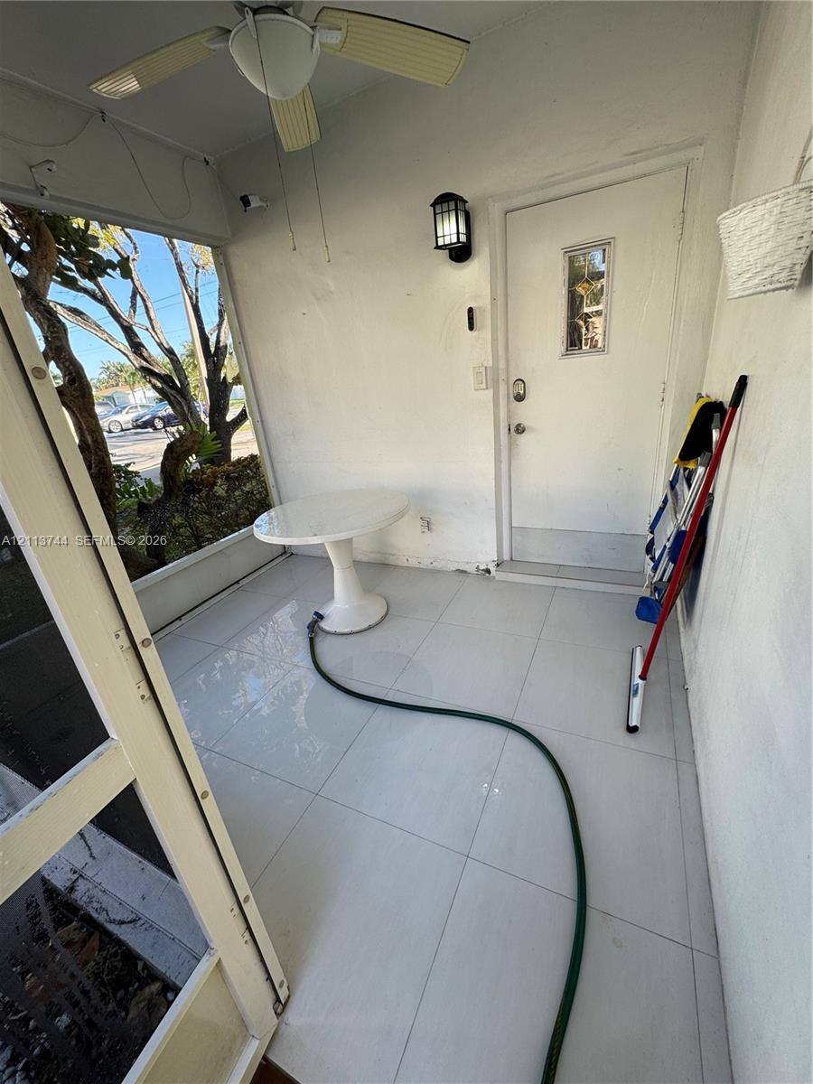 Enclosed porch featuring large-format tile flooring, a ceiling fan with integrated lighting, a wall-mounted lantern-style sconce, and a front entry door with a decorative glass insert
