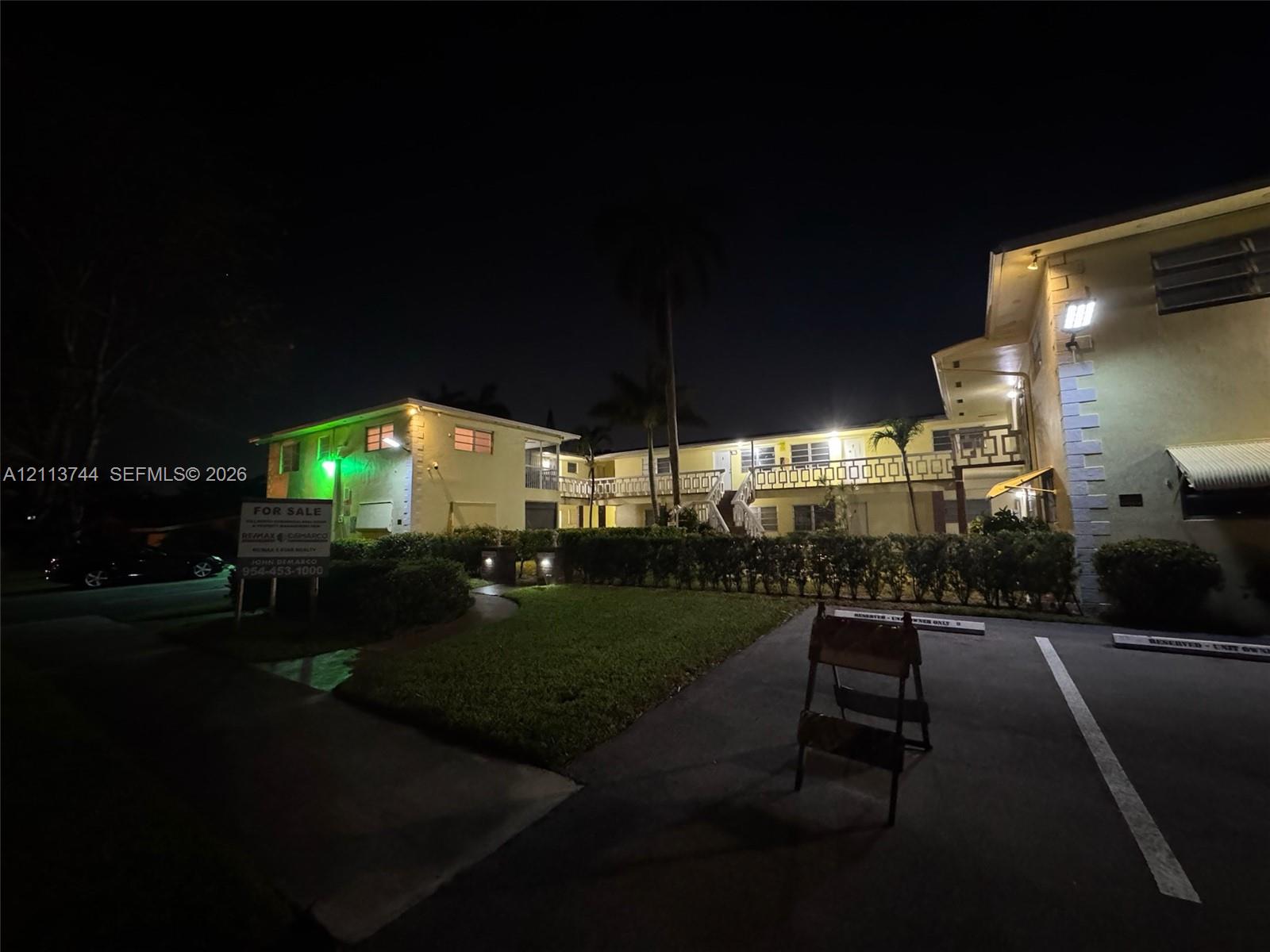 Multi-story residential building featuring a stucco exterior, second-level balcony with decorative metal railing, manicured lawn, and dedicated asphalt parking area